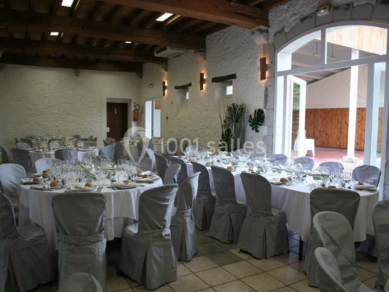 Salle de réception avec tables rondes dressées, nappes blanches et chaises couvertes, éclairée par des ouvertures vitrées.