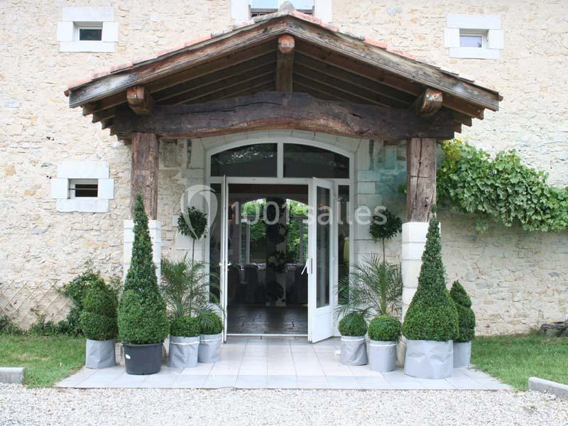 Entrée d'un bâtiment en pierre avec porte vitrée, encadrée de plantes en pots et d'un auvent en bois.