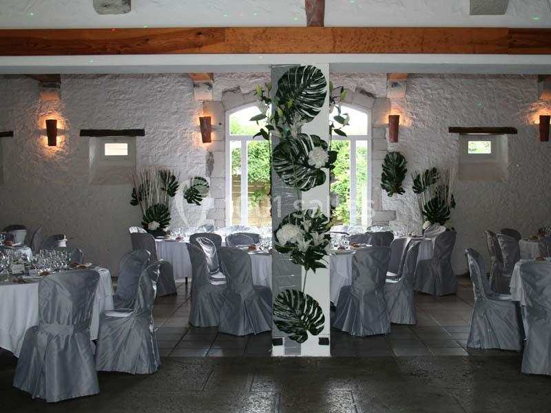 Salle de réception décorée avec des nappes et housses de chaises grises, ornée de feuillages tropicaux et de fleurs blanches.