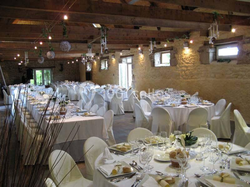 Salle de réception rustique avec tables décorées, nappes blanches, chaises houssées et lumière naturelle.