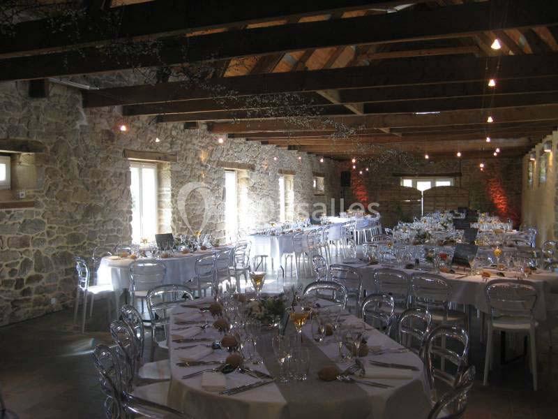 Salle en pierre avec poutres apparentes, tables rondes dressées pour un événement, chaises transparentes et lumière…