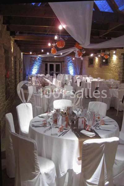 Salle de réception décorée avec des tables rondes, nappes blanches, chaises houssées et éclairage tamisé.