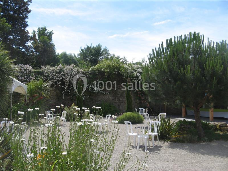 Jardin avec des fleurs blanches, des chaises en plastique dispersées et des arbres sous un ciel partiellement dégagé.