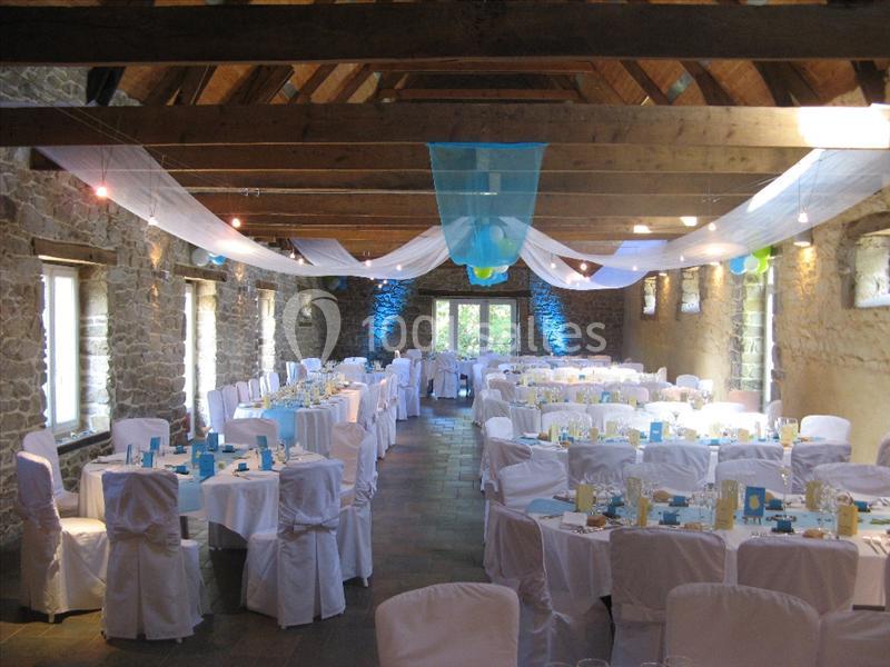 Salle en pierre avec poutres apparentes, décorée pour un événement, tables dressées avec nappes blanches et voilages…