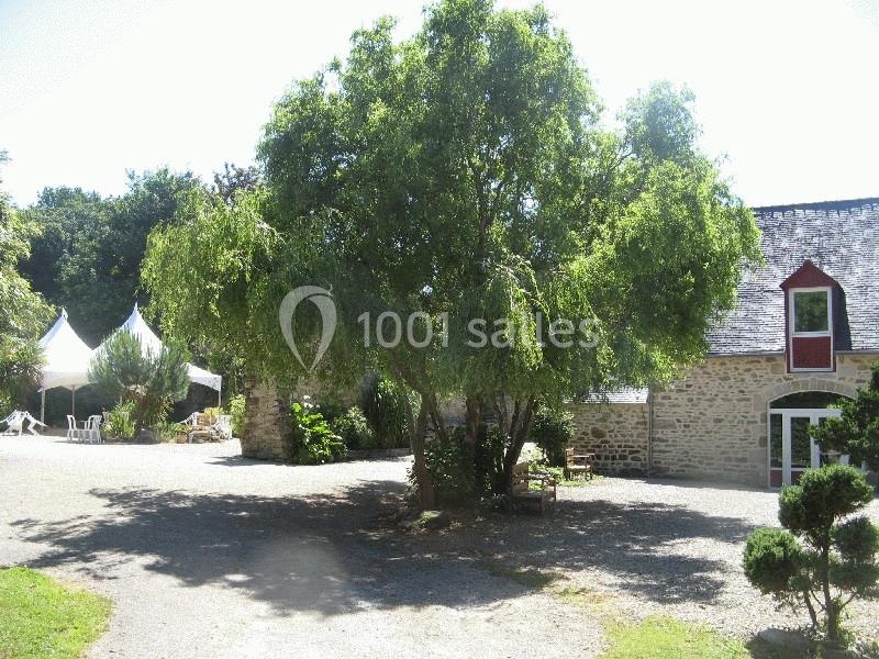 Cour avec un grand arbre au centre, entourée de bâtiments en pierre et de tentes blanches en arrière-plan.