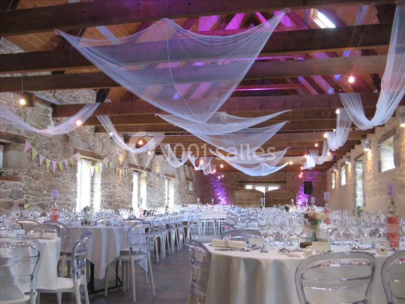 Salle de réception en pierre avec tables dressées, chaises transparentes et voilages suspendus au plafond en bois.
