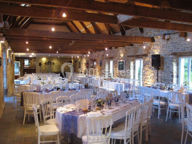 Salle de réception en pierre avec poutres apparentes, tables dressées et chaises blanches pour un événement.