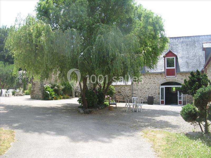Cour avec un grand arbre, des chaises blanches et une maison en pierre avec une porte et des fenêtres rouges.