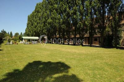 Grand jardin avec pelouse, arbres alignés et une structure couverte avec des tables et chaises blanches.