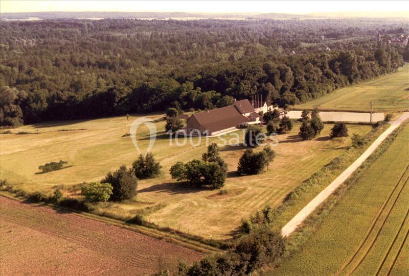 Vue aérienne d'une ferme entourée de champs et d'arbres, avec une forêt en arrière-plan.