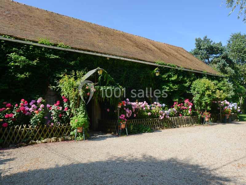 Façade d'une grange en pierre recouverte de végétation, entourée de rosiers fleuris et d'un chemin gravillonné.