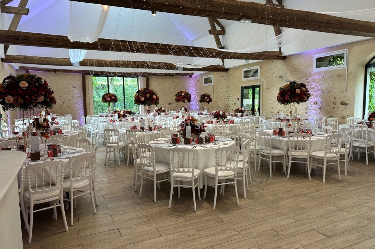 Salle de réception décorée avec des tables rondes, nappes blanches, chaises assorties et centres de table fleuris.