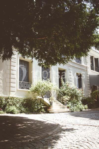 Façade d'une maison blanche avec volets bleus, entourée de végétation et donnant sur une allée pavée.