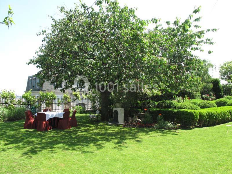 Table dressée sous un grand arbre dans un jardin verdoyant, entourée de buissons taillés et de fleurs.