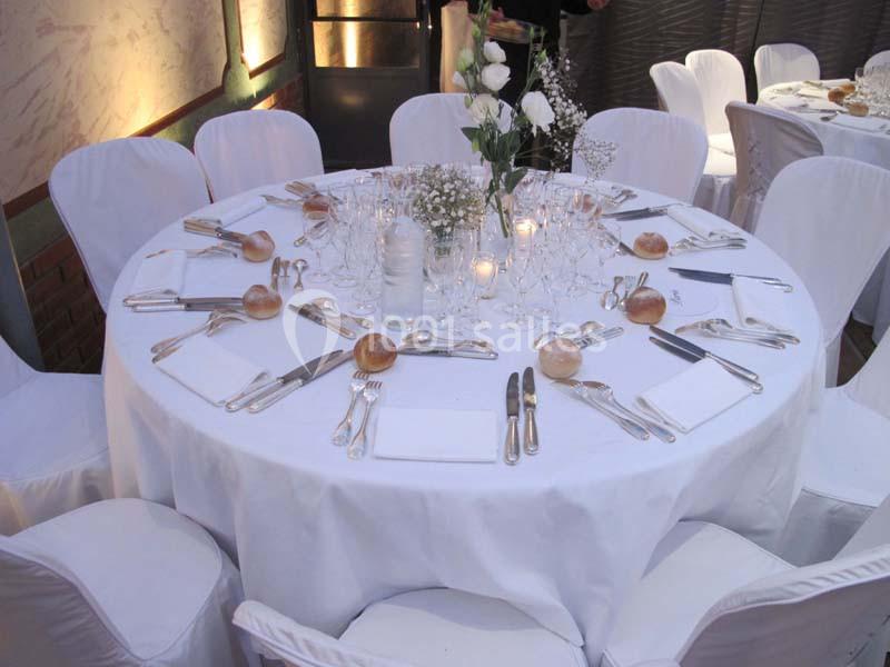 Table ronde dressée pour un repas, avec nappes blanches, couverts, verres, fleurs et petits pains disposés.