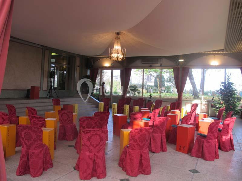 Chaises recouvertes de housses rouges et tabourets colorés disposés dans une salle lumineuse avec lustre et grandes fenêtres.