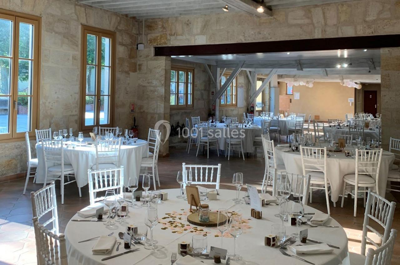 Salle de réception lumineuse avec tables rondes dressées, chaises blanches et murs en pierre apparente.