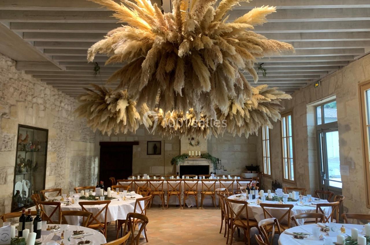 Salle de réception avec tables dressées et grande suspension décorative en herbes séchées au plafond.
