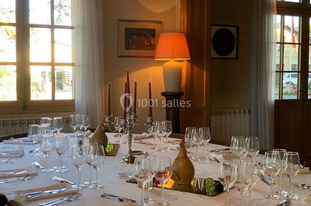 Table dressée avec nappes blanches, verres, bougeoirs et décorations, dans une salle lumineuse avec fenêtres et lampes.