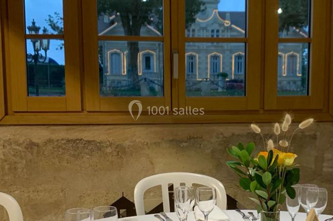 Table dressée avec vaisselle et verres, décorée d'un bouquet, devant une fenêtre donnant sur un bâtiment éclairé.