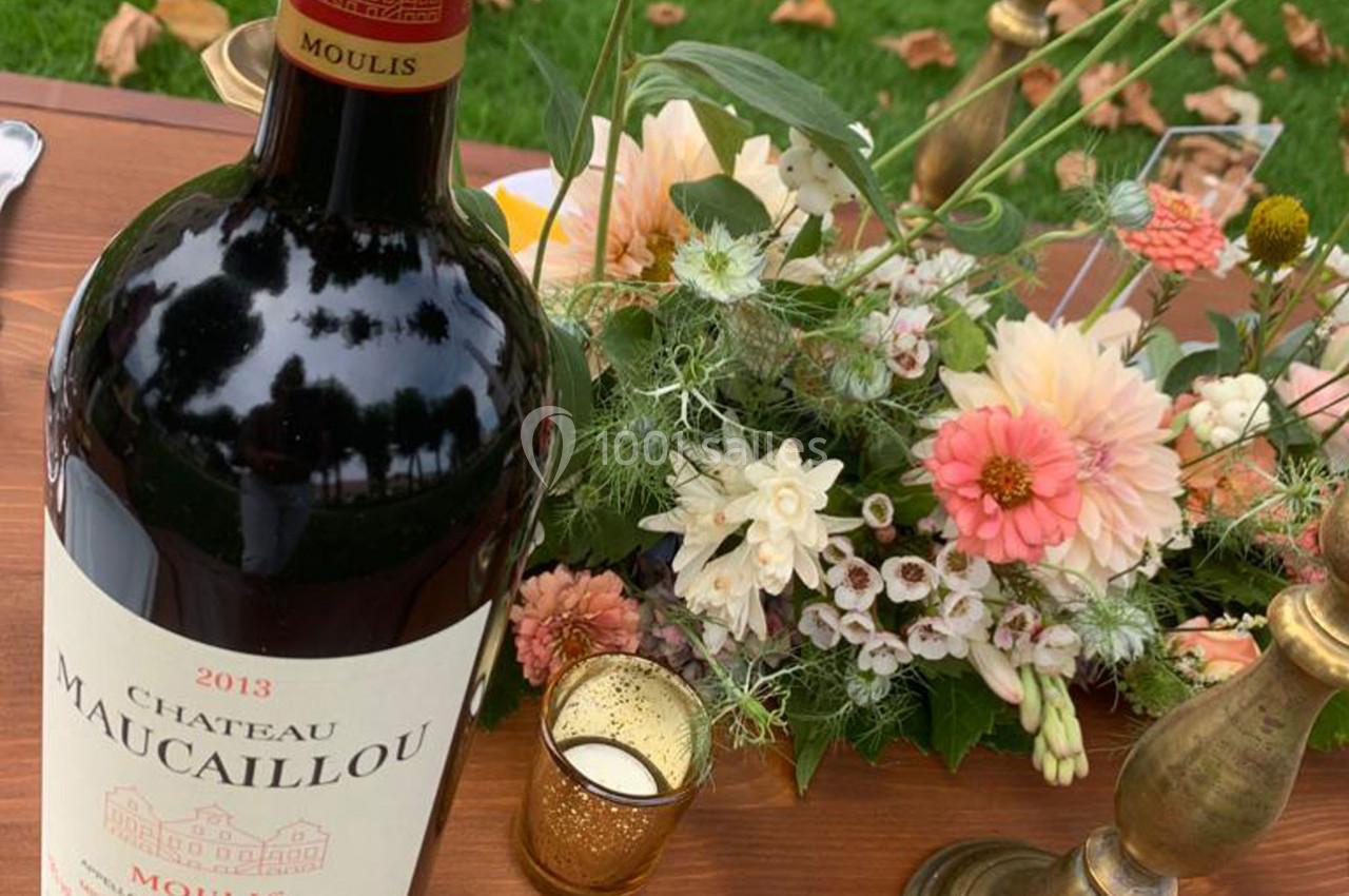 Bouteille de vin rouge Château Maucaillou 2013 posée sur une table en bois avec un bouquet de fleurs et des bougeoirs.