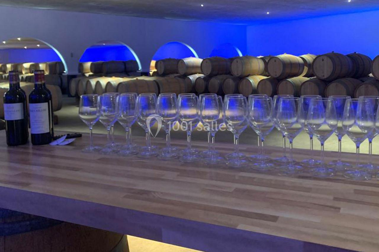 Rangée de verres à vin vides sur un comptoir en bois, avec des barriques et deux bouteilles en arrière-plan.