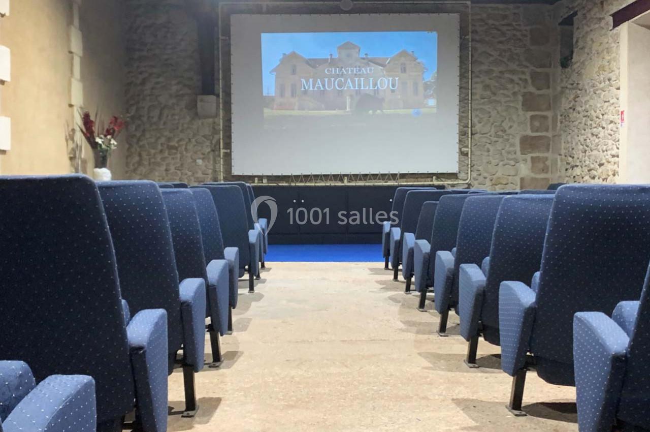 Salle de projection avec des rangées de fauteuils bleus et un écran affichant une image du Château Maucaillou.