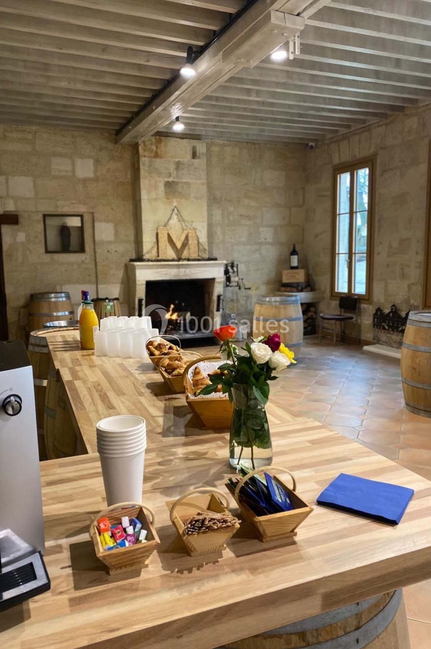 Salle rustique avec cheminée, tables en bois, barriques, et buffet comprenant viennoiseries, boissons et fleurs.