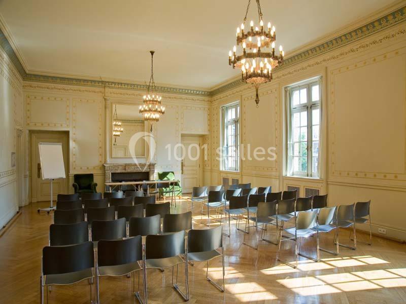 Salle lumineuse avec parquet, chaises alignées, tableau de conférence, cheminée et lustres suspendus.