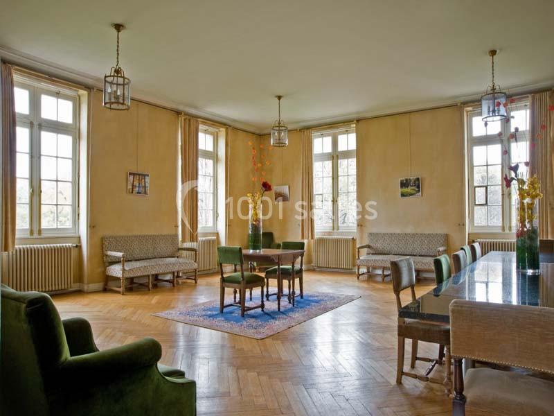 Salle lumineuse avec parquet en bois, mobilier ancien, grandes fenêtres et décoration florale sur une table centrale.