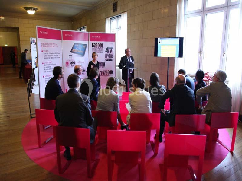 Un groupe de personnes assises sur des chaises rouges écoute une présentation dans une salle lumineuse.