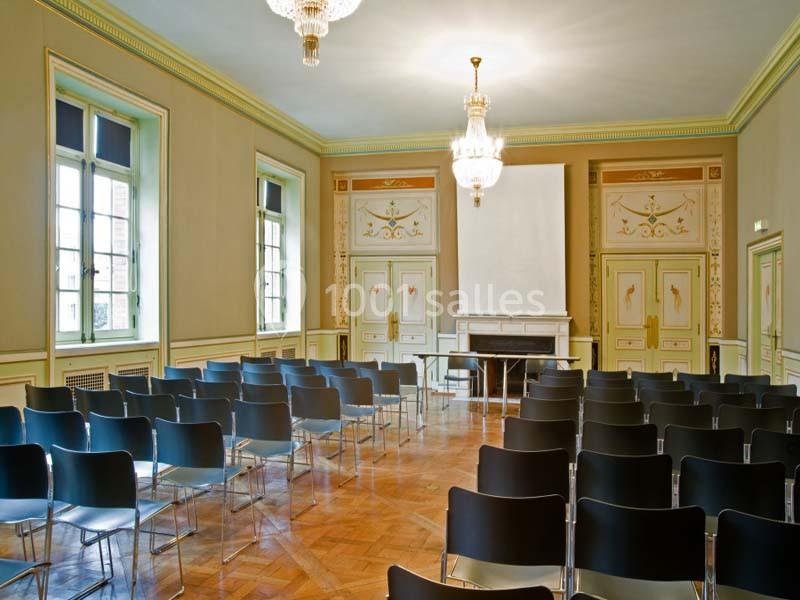 Salle lumineuse avec parquet, chaises noires alignées, cheminée, lustres et murs décorés de motifs classiques.