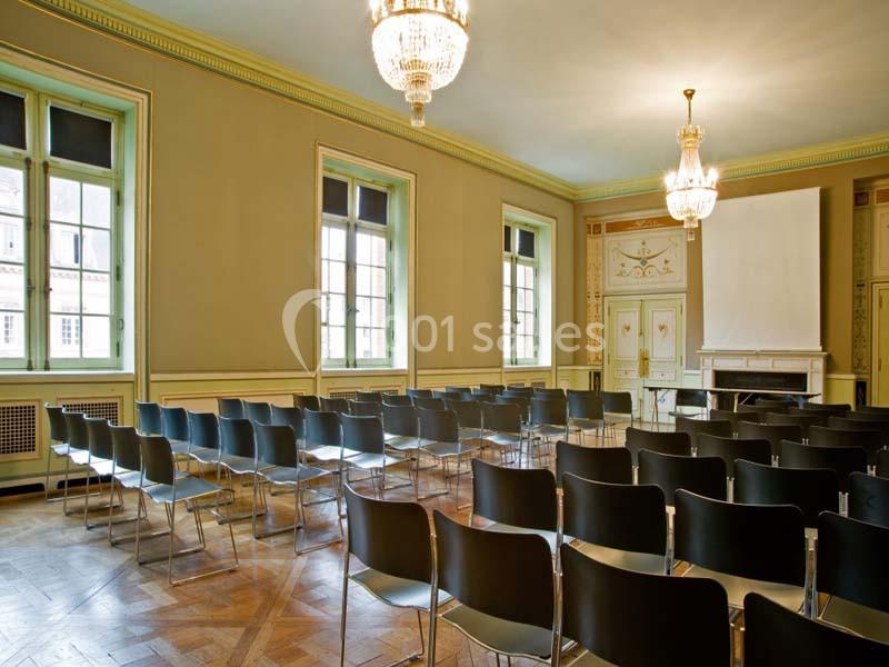 Salle de conférence lumineuse avec rangées de chaises noires, parquet en bois et grand écran blanc devant.