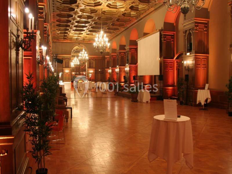 Grande salle élégante avec parquet, hauts plafonds ornés, lustres et tables rondes dressées pour un événement.