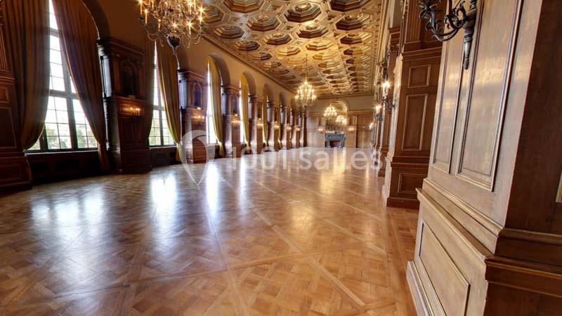 Grande salle avec parquet en bois, hauts plafonds ornés, lustres suspendus et grandes fenêtres laissant entrer la lumière.