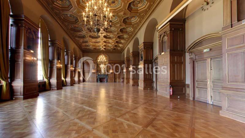 Grande salle avec parquet en bois, hauts plafonds ornés, lustres imposants et murs décorés de boiseries.