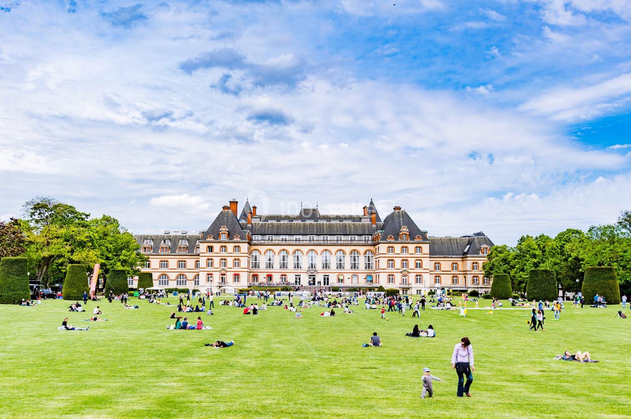 Pelouse avec des personnes assises ou allongées devant un grand bâtiment historique entouré d'arbres.