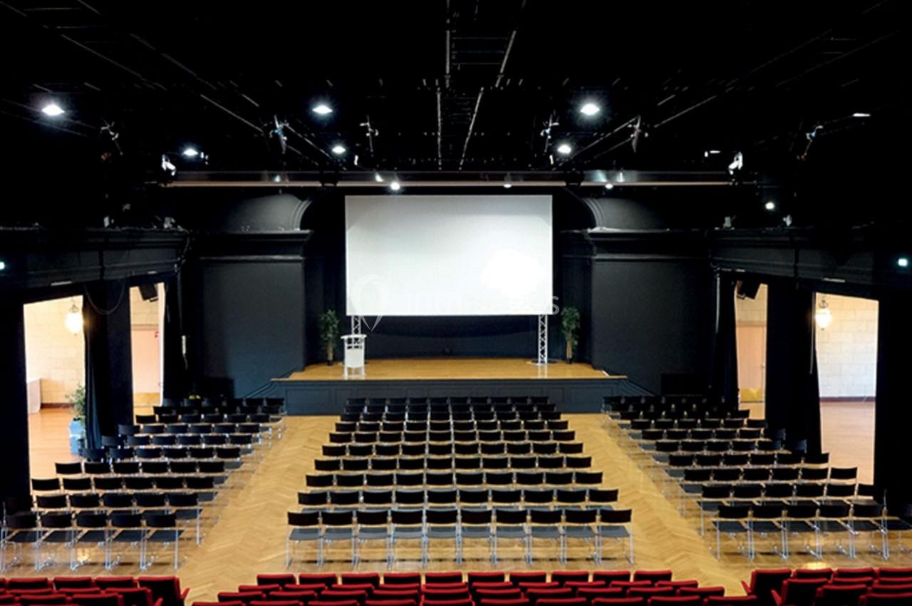 Salle de conférence avec un écran géant, des rangées de chaises noires et un pupitre sur une scène éclairée.