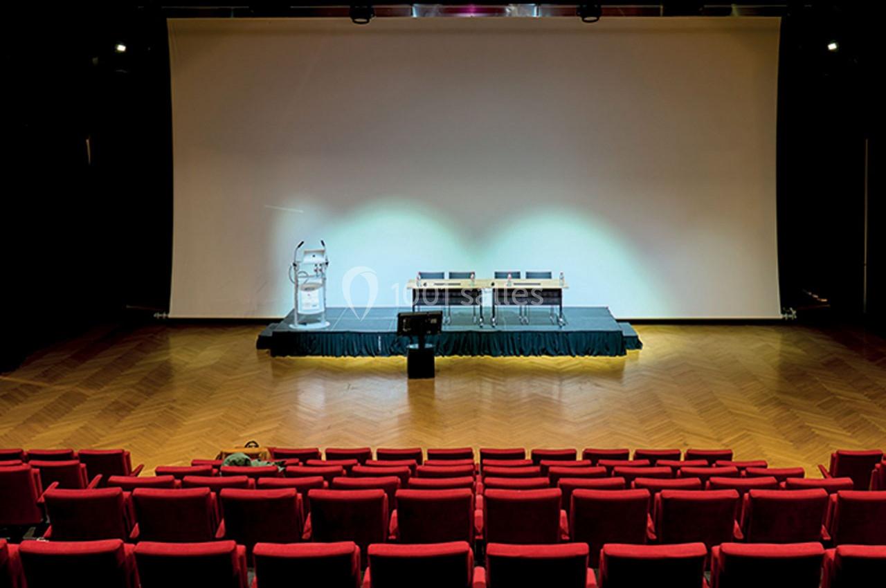 Salle de conférence vide avec rangées de sièges rouges, scène équipée d'un pupitre, d'une table et d'écrans.
