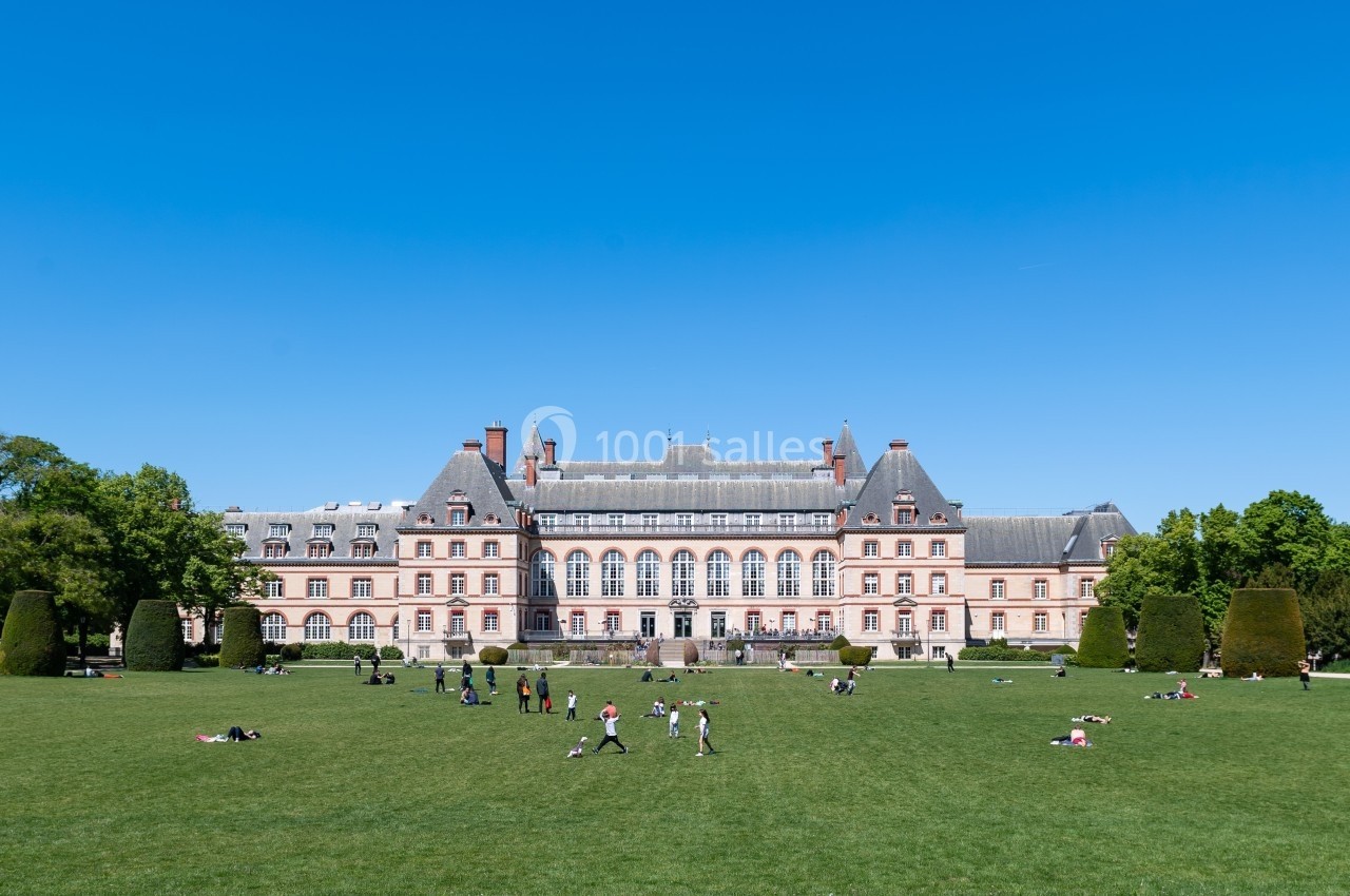 Façade d'un grand bâtiment historique avec un toit mansardé, entouré d'une pelouse où des personnes se détendent.