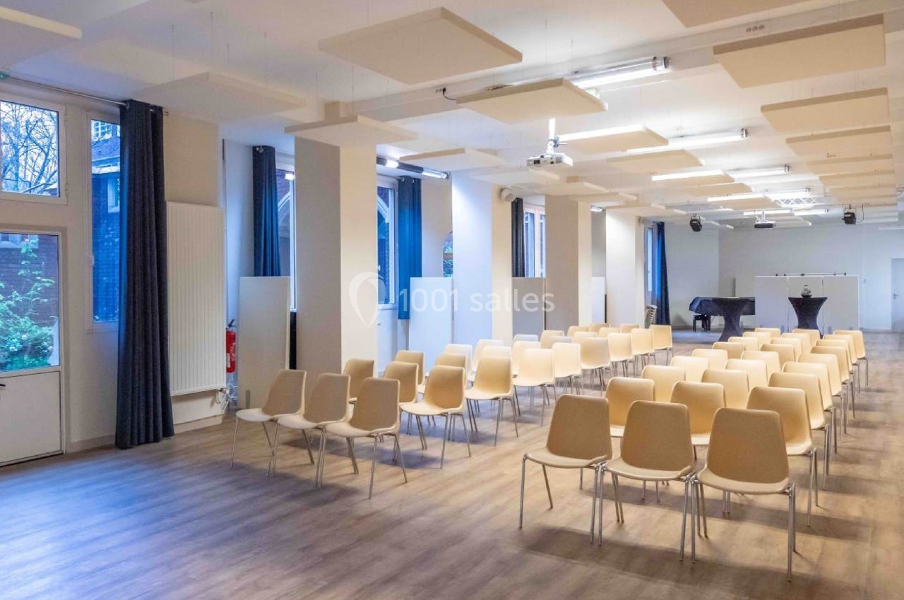 Salle lumineuse avec des rangées de chaises beige, un piano noir au fond et des panneaux acoustiques au plafond.