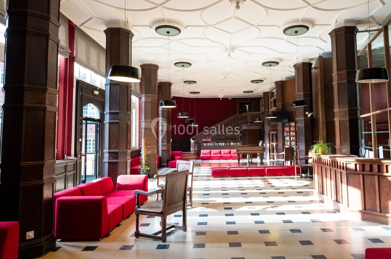 Salle lumineuse avec colonnes en bois, canapés rouges, sol en damier et escalier menant à une mezzanine.