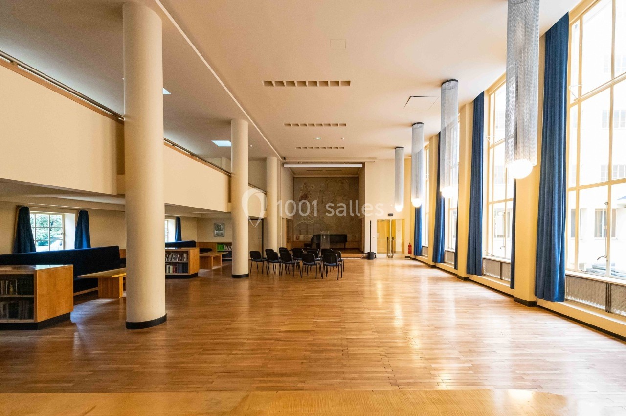 Salle spacieuse avec parquet, colonnes blanches, grandes fenêtres et chaises alignées près d'un mur décoré.