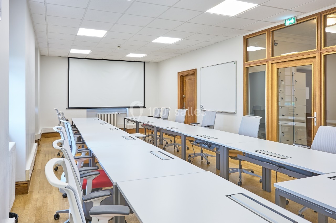 Salle de réunion lumineuse avec de longues tables blanches, des chaises modernes et un écran de projection.