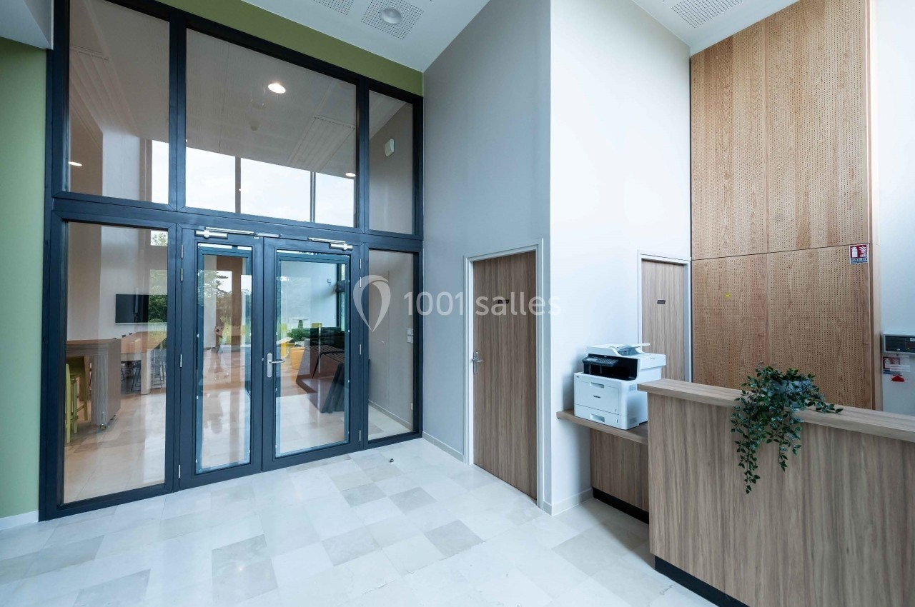 Entrée d'un bâtiment avec portes vitrées, comptoir d'accueil en bois, imprimante et plante décorative.