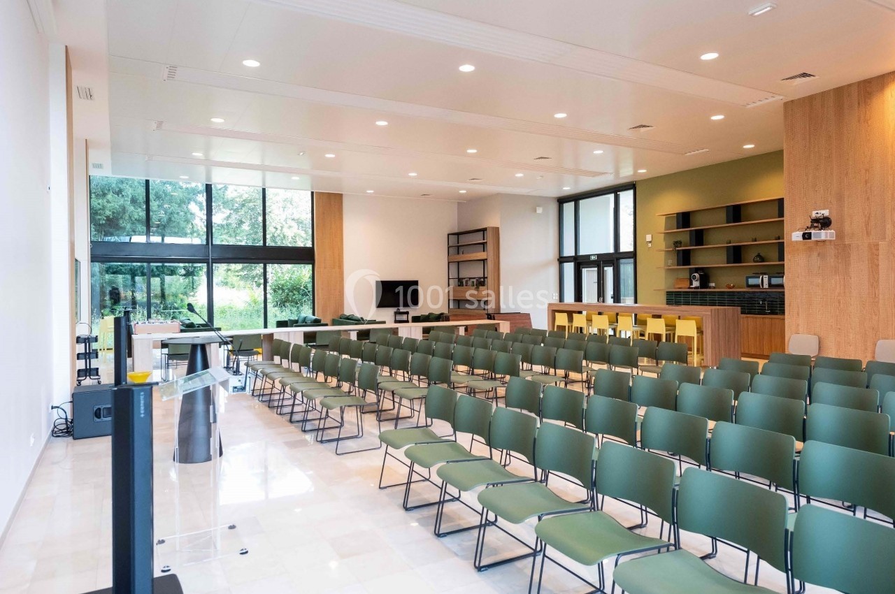 Salle lumineuse avec rangées de chaises vertes, pupitre à gauche, bar et étagères en bois au fond, grandes baies vitrées.