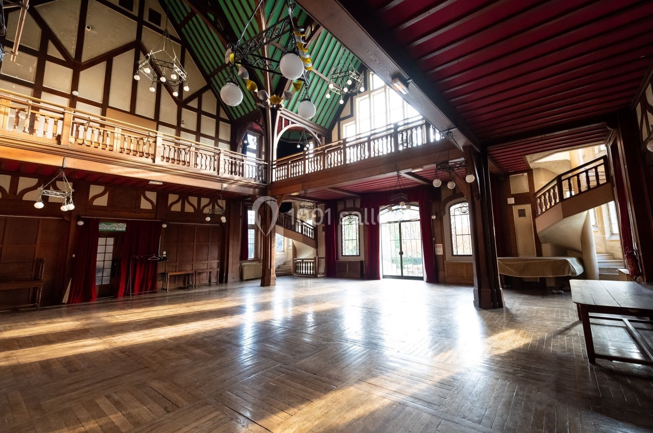 Grande salle lumineuse avec parquet en bois, balustrades, poutres apparentes et plafond coloré.