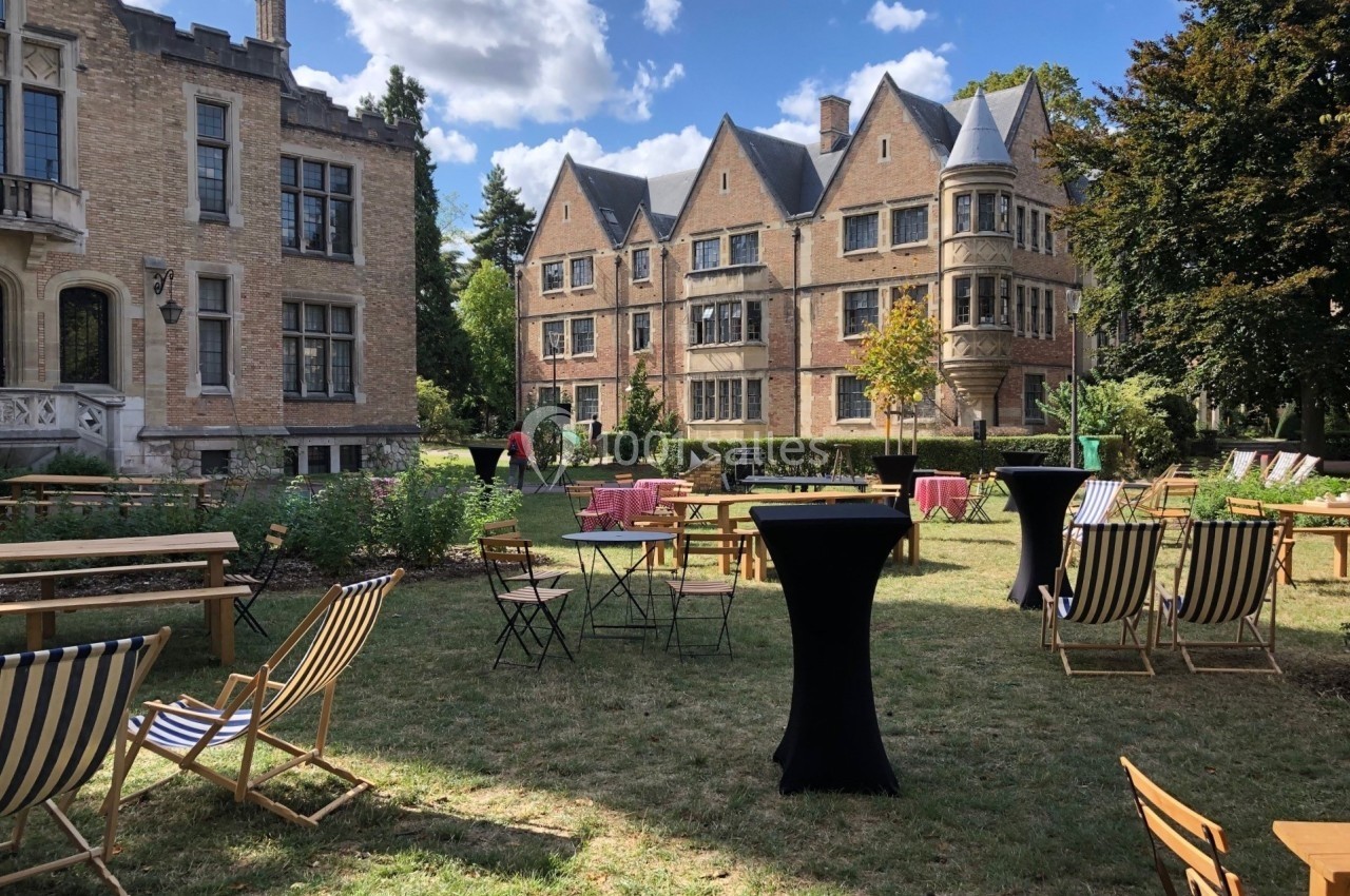 Jardin avec chaises longues, tables hautes et bâtiments en briques dans un cadre ensoleillé et verdoyant.