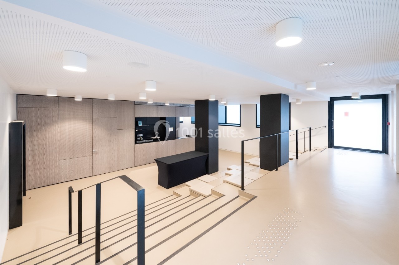 Espace intérieur moderne avec escalier, comptoir d'accueil, éclairage suspendu et sol clair.