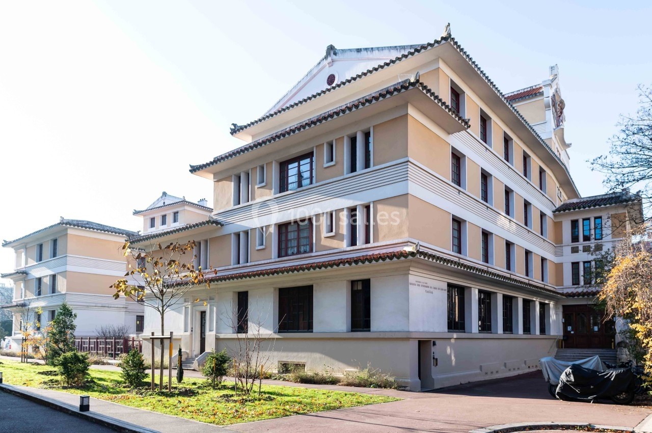 Bâtiment de style asiatique avec plusieurs étages, toits en pagode et façade beige, entouré d'arbres et d'un trottoir.